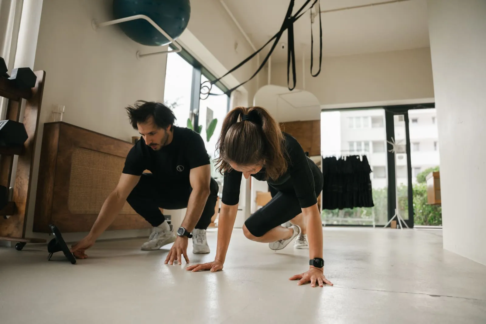A coach spotting a client in an EMS suit during a plank exercise by the window