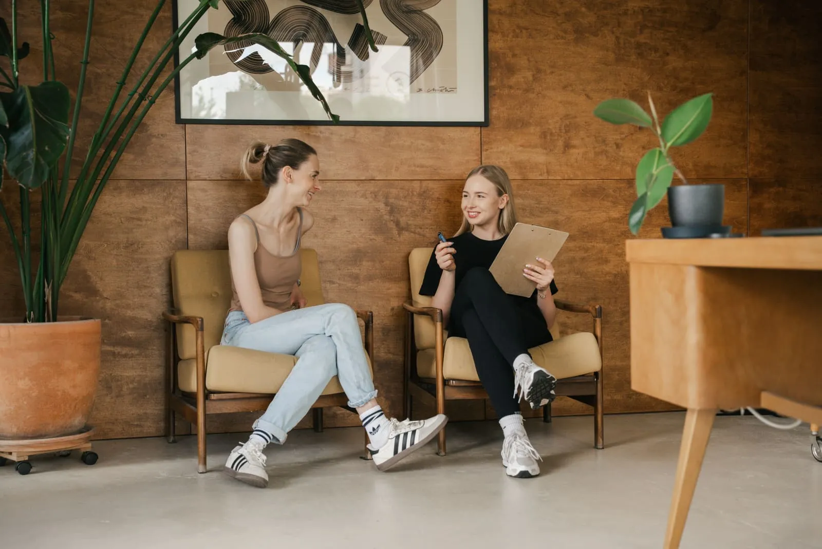 Two clients talking in armchairs by the wooden wall, one holding an intake form