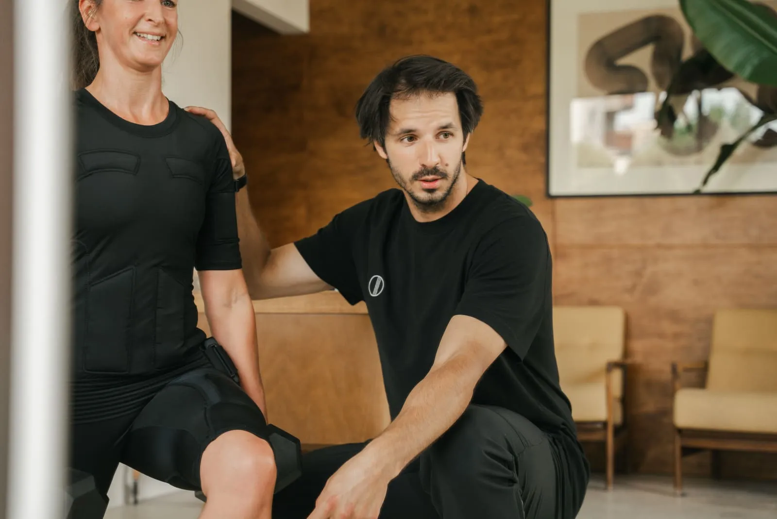 A client in an EMS suit and a coach sitting by the studio's wooden wall