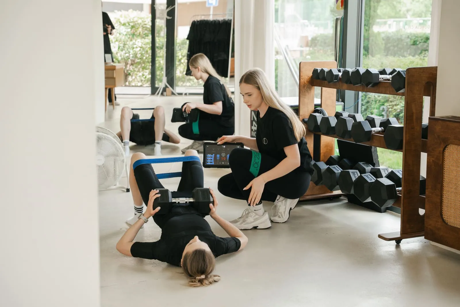 A coach guiding a client through a mat exercise, a dumbbell rack in the background
