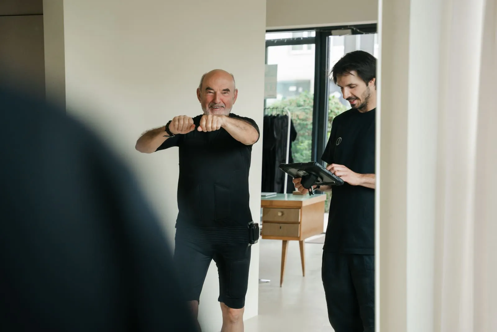 An older client in an EMS suit performing an exercise while the coach monitors the session
