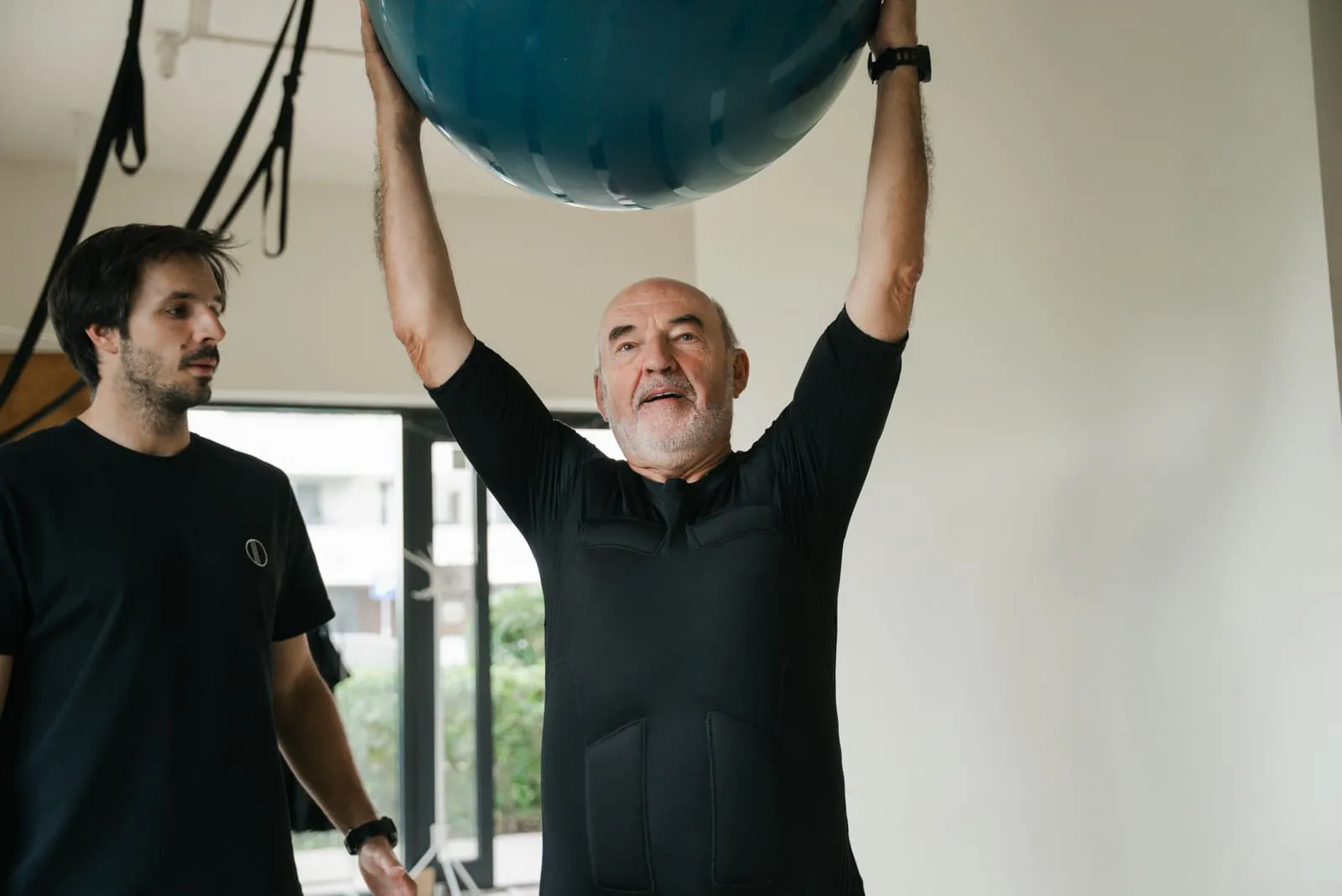 An older client raising an exercise ball overhead under a coach's eye