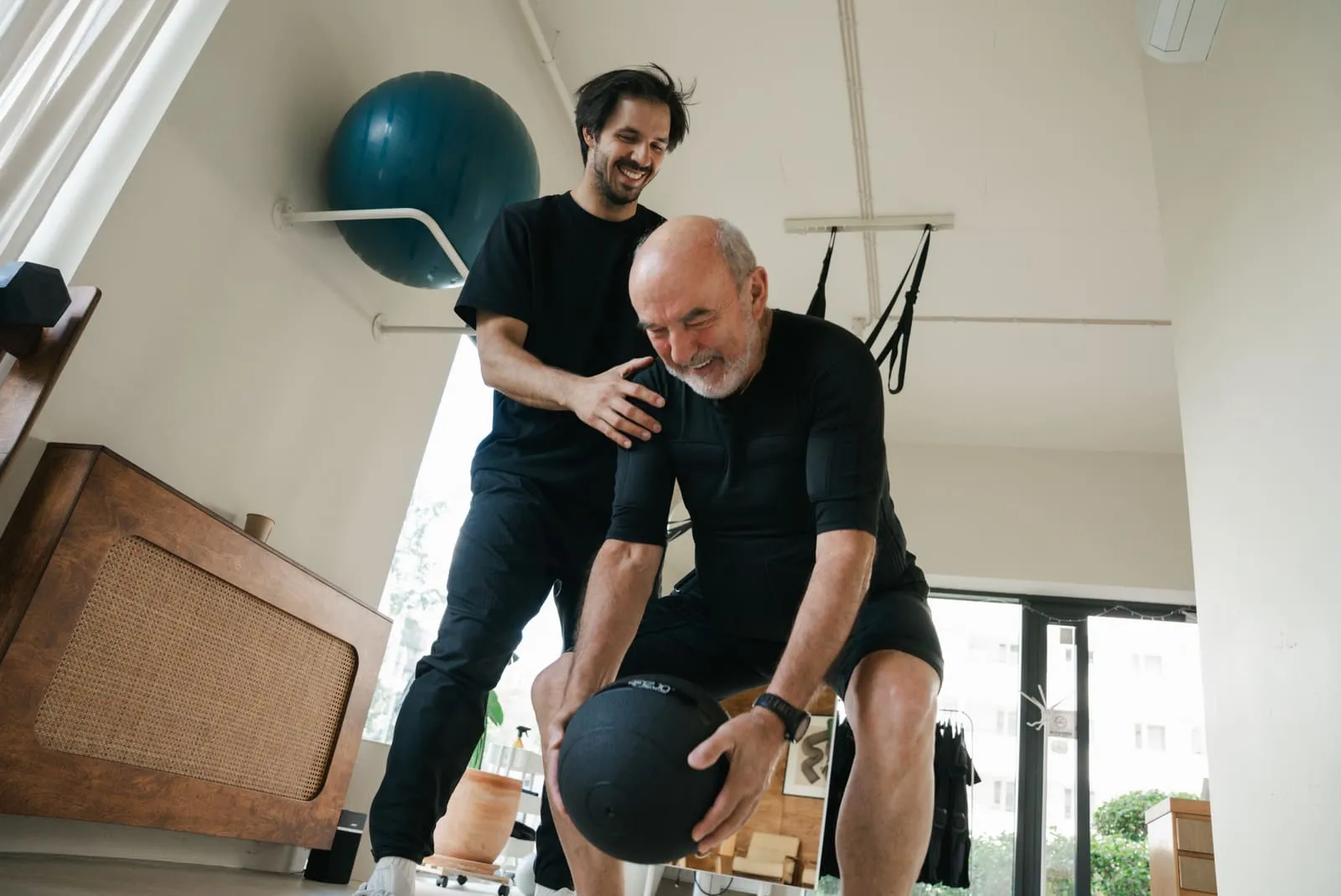 An older client training with a medicine ball, the coach spotting by the window