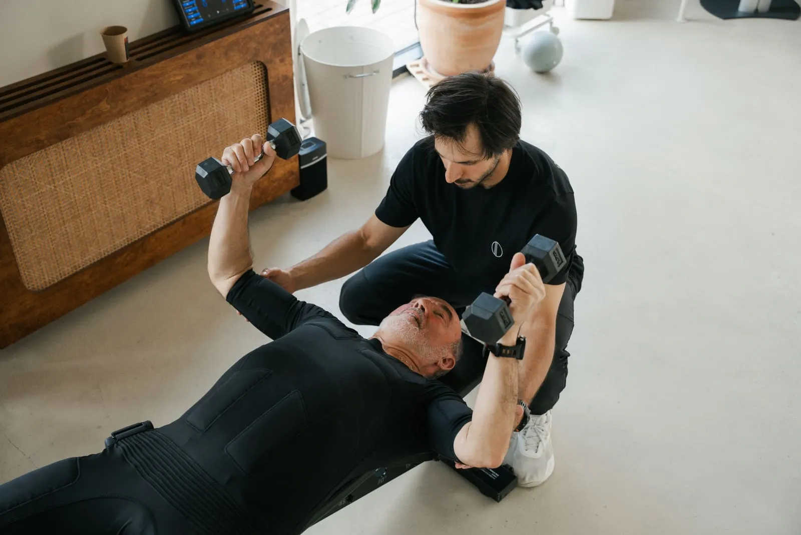 A coach handing dumbbells to a client in an EMS suit lying on the mat
