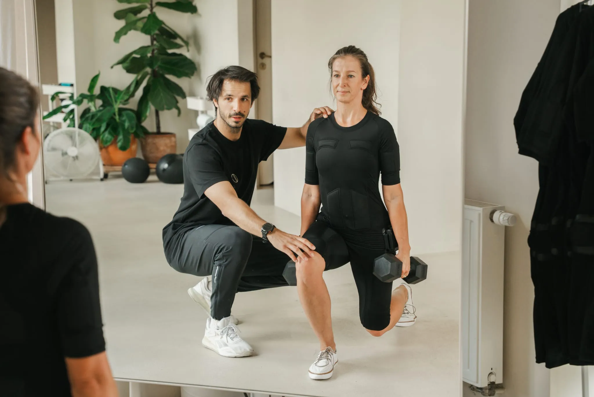 A coach spotting a client in an EMS suit during a dumbbell lunge, seen from the mirror
