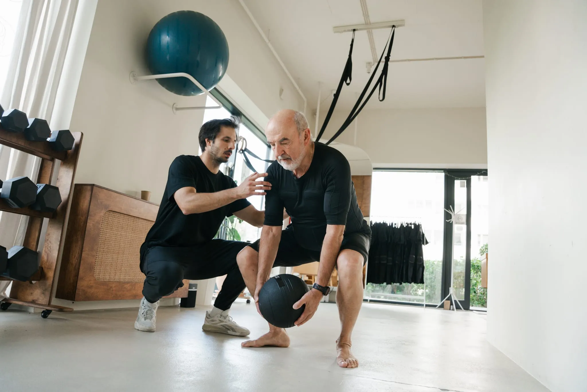 An older client in an EMS suit lifting a medicine ball off the floor, the coach crouching beside him