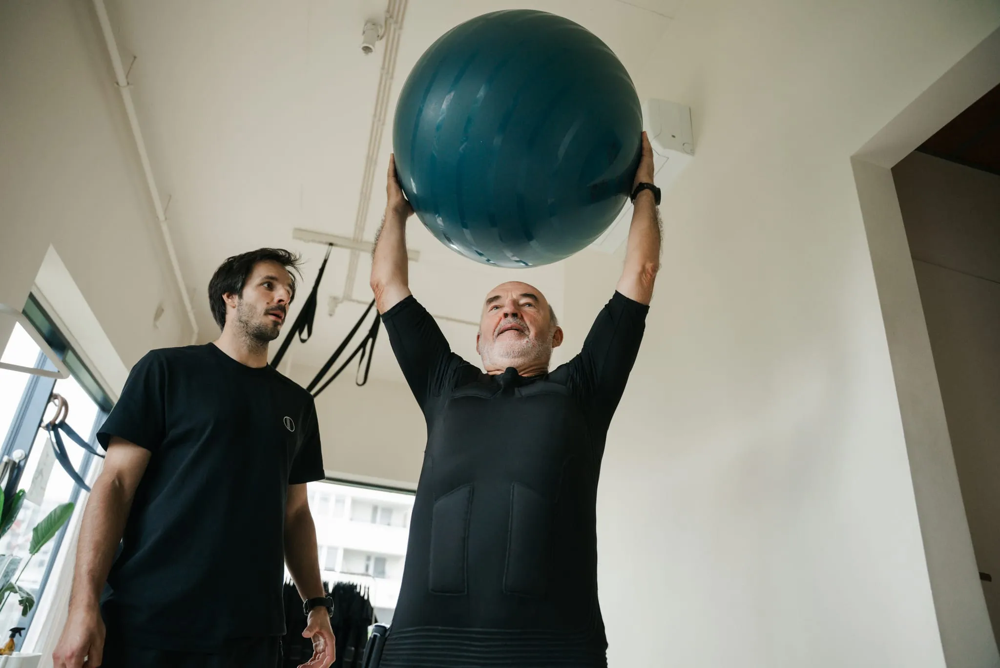 An older client in an EMS suit pressing a large ball overhead under a coach's eye