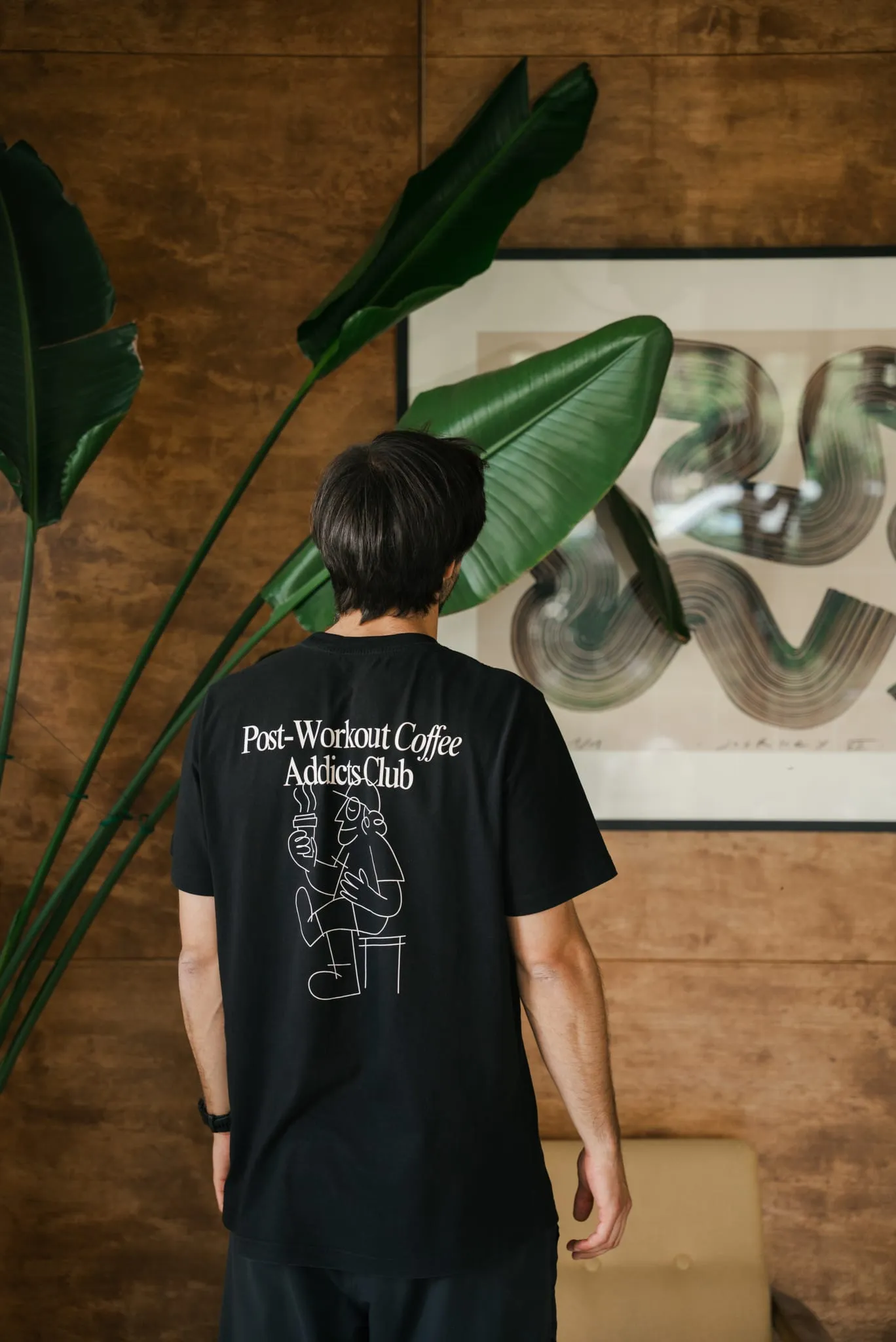 Mężczyzna w T-shircie WORK IT przed drewnianą ścianą studia obok rośliny i grafiki