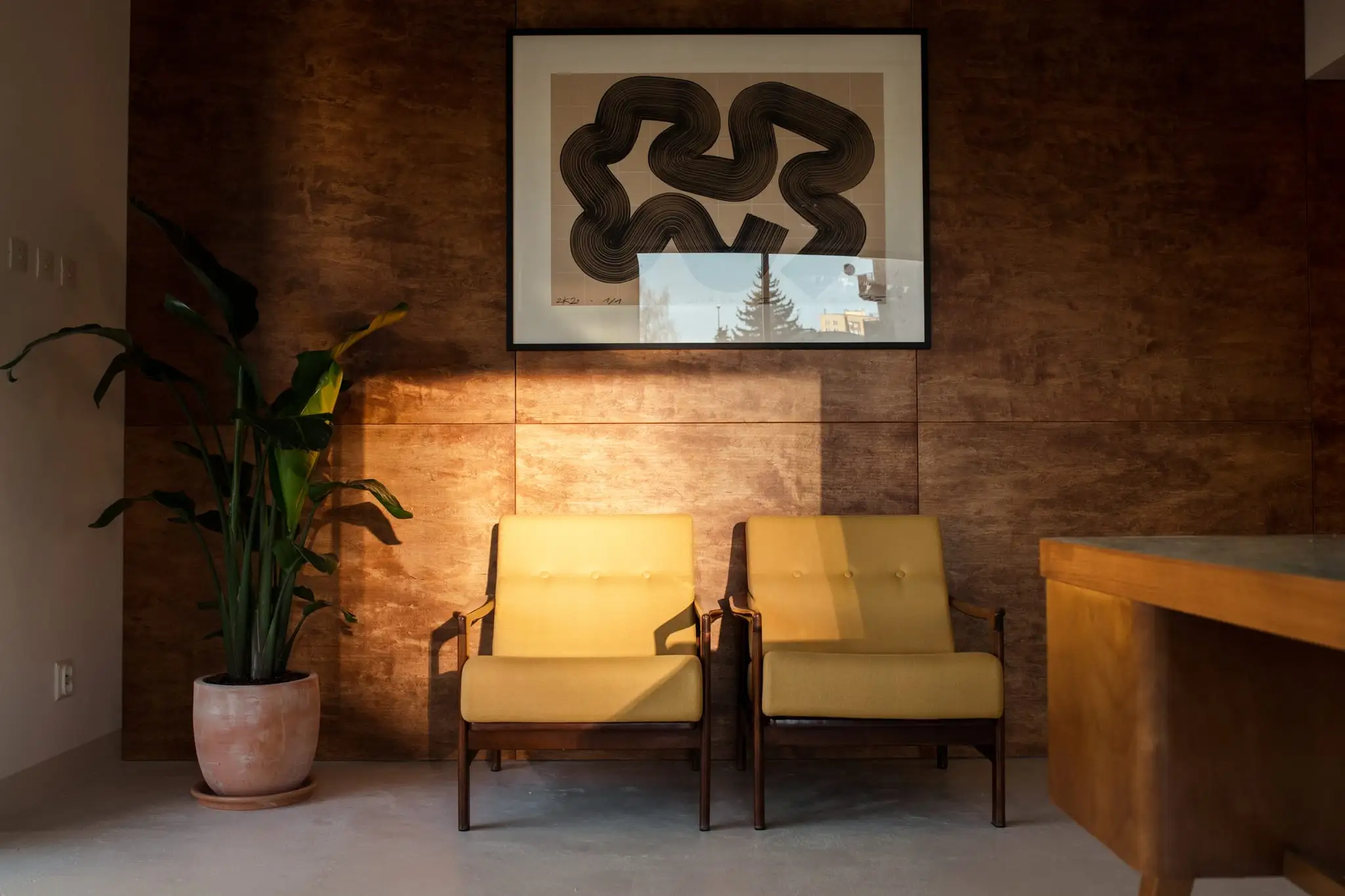 Two yellow armchairs beneath artwork on the studio's wooden wall, a potted plant beside them in a shaft of sunlight